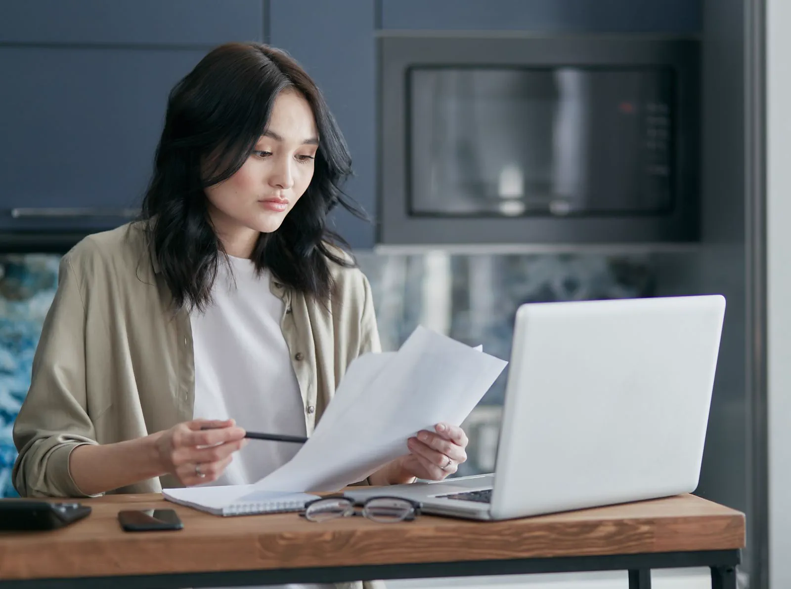Woman reading papers at the kitchen table, with a laptop in front of her